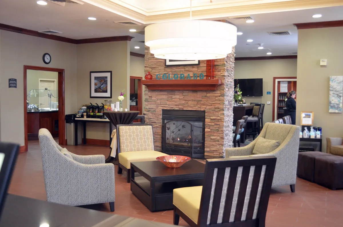 A cozy hotel lounge featuring a welcoming fireplace, a comfortable seating arrangement with plush chairs and sofas, a stylish coffee table, and warm lighting. Guests are seen relaxing, socializing, and enjoying refreshments together, creating an inviting, communal atmosphere.