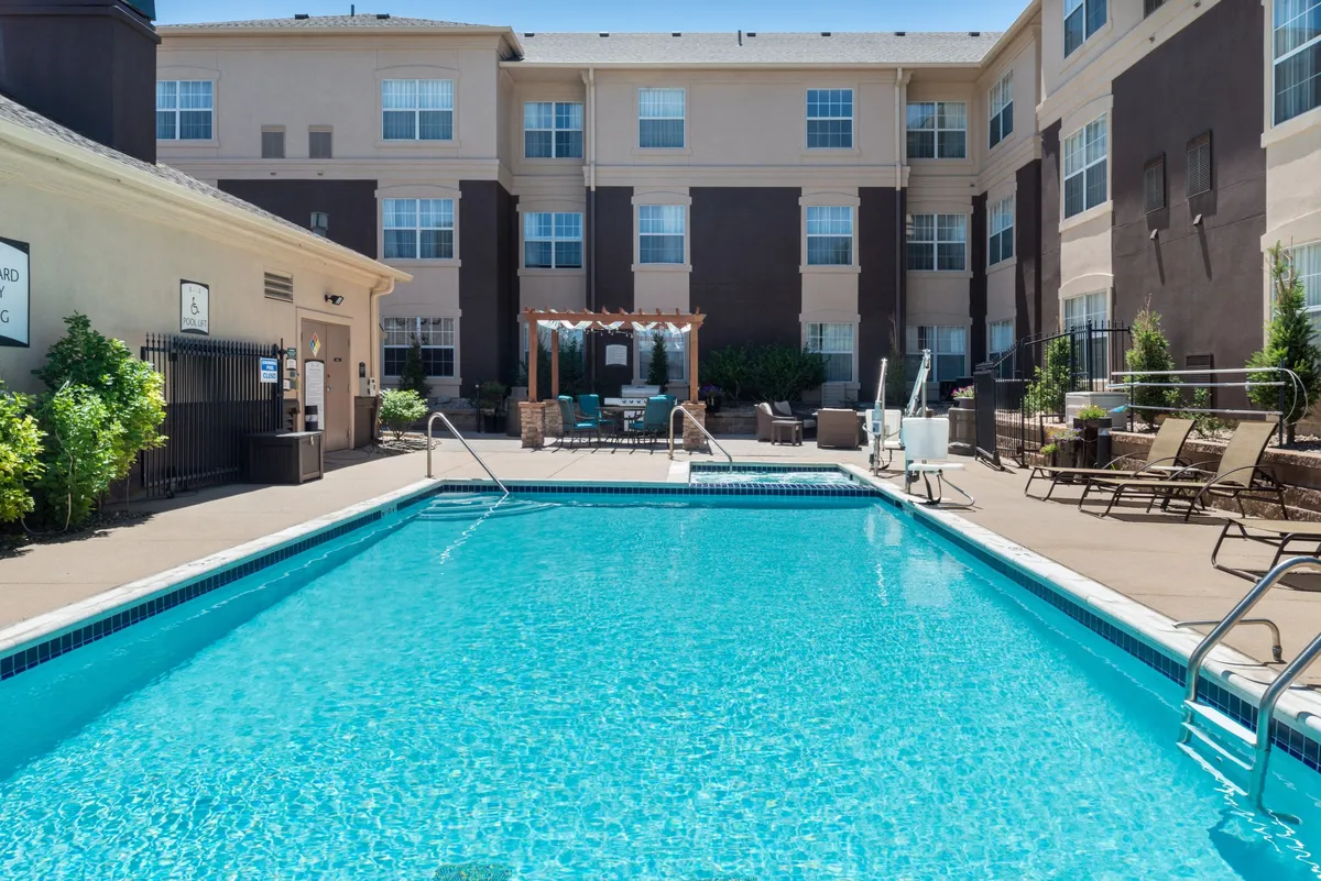 This image features a sparkling outdoor swimming pool surrounded by comfortable lounge chairs and a shaded seating area under a pergola. Lush landscaping provides a welcoming atmosphere, and the setting sun casts a warm, inviting glow over the space. The area suggests both relaxation and opportunities for socializing, with multiple spots to unwind or gather with others.