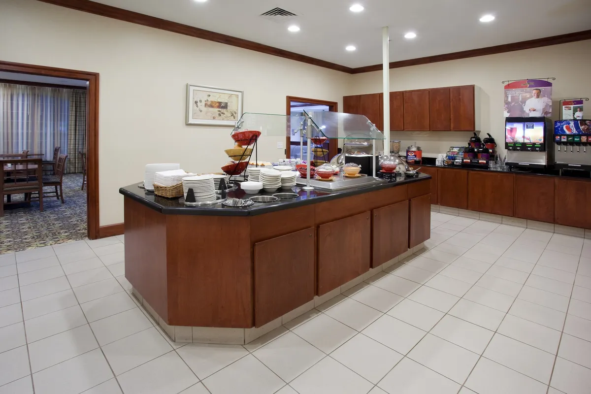 A bright, modern breakfast area featuring a well-stocked breakfast bar with a variety of food and beverage options. The space is inviting, with several guests enjoying breakfast and engaging in conversation, evoking a communal and energetic start to the day.