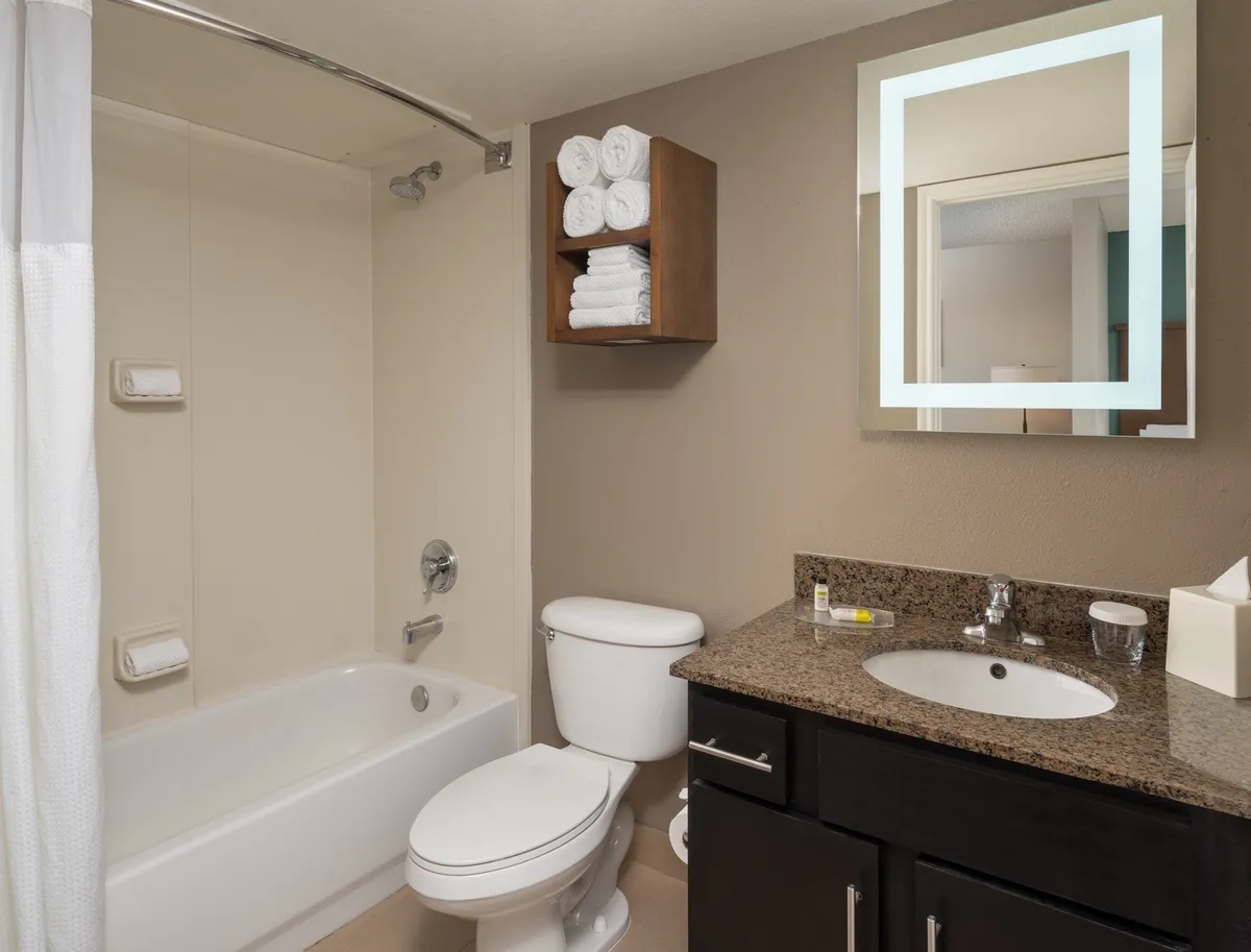 This image depicts a modern bathroom with a bathtub, sink, large mirror, and neatly arranged fresh towels. The space exudes cleanliness and functionality, highlighting amenities that contribute to a hotel-like experience. The mood is inviting and functional, suggesting a comfortable stay with thoughtful provisions.