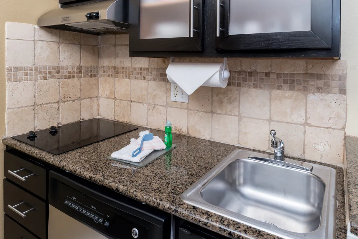 The image showcases a stylish, modern kitchen with sleek black cabinets, a polished granite countertop, stainless steel sink, and a chic tile backsplash. The space is well-lit and feels both inviting and functional, perfect for meal prep or gathering around for a casual bite.