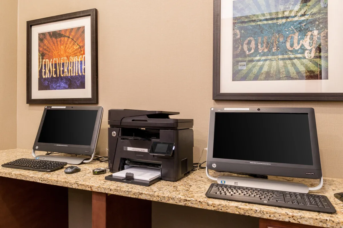 A sleek, modern business center featuring desktop computers, a printer, and motivational artwork on the walls. The space is well-lit and organized, ideal for focused work sessions. The atmosphere conveys productivity and professionalism, catering specifically to business travelers and remote workers.