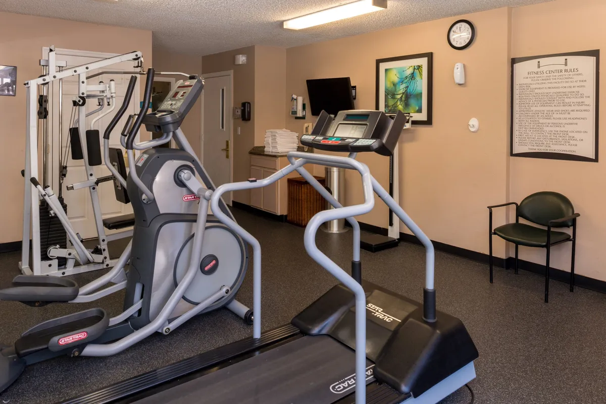 A vibrant, well-lit fitness center featuring treadmills, elliptical machines, strength training equipment, and a fitness rules poster. The space is inviting for guests looking to decompress after a workday or fit in a workout between meetings.