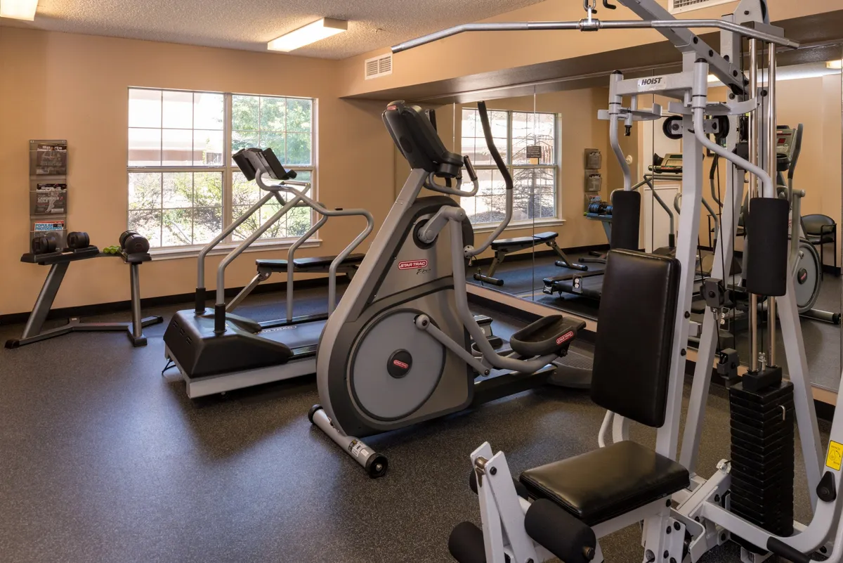 A modern fitness room equipped with an elliptical machine, stationary bike, weight machine, a mirrored wall, and windows that bring in natural light. The space is energized, suggesting guests can keep active during their stay.
